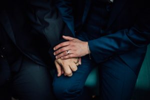 Close up shot of hands of two grooms wearing wedding rings at a theatre wedding. Hands resting on their knees with dark blue suits showing.