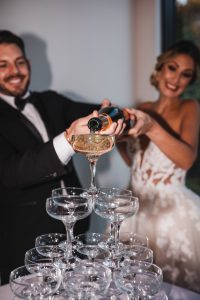 Bride and Groom laughing whilst pouring champagne in to a glass tower. A wedding trend 2023