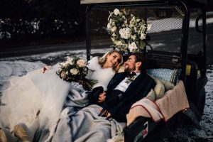 Bride and groom laying on the back of a golf buggy. There is snow and frost behind on the floor and the sun is hitting their faces.