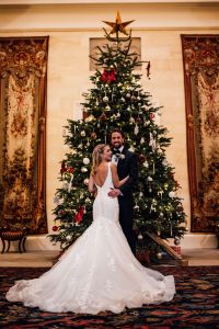 Bride and groom stand in front of a giant Christmas tree in the entrance to Kinmount House. Brides dress is fanned out being her.