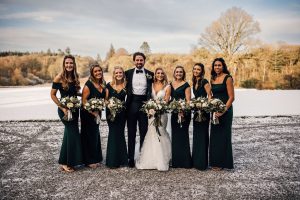 Bride and groom surrounded by their bridesmaids who are in dark green floor length dresses holding bouquets. Snow covered field behind them. 