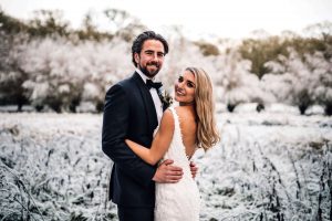 Bride and groom holding each other in an embrace facing the camera both smiling. There are snow covered trees in the back ground and thick frost on the ground. Taken in the early evening at Kinmount House.