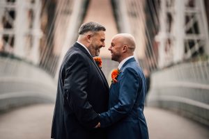 Two grooms standing facing each other, holding hands. Matching blue suits with an orange button hole.