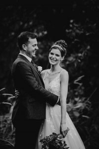 Black and white photo of a bride and groom holding each other around the waist. The groom looks at the bride and the bride looks at the camera.