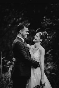 Bride and groom holding each other around the waist. They are both smiling as if about to burst out laughing. She hods flowers in her left hand he wears a button hole. Black and white image.