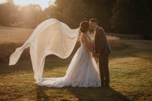 Bride and groom kissing at sunset. The brides veil is blowing behind her and the sun is setting behind them. Standing in a field at Bassmead Manor Barns. 