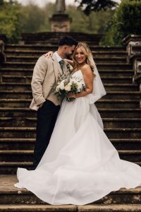 Bride and groom on the steps of harlaxton Manor. The groom has his head resting on the brides head as she looks to the camera.