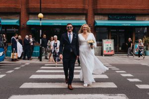 Colour image of a bride and groom walking across a zebra crossing on their way to a boat party wedding. In the back ground you can see their guests walking.