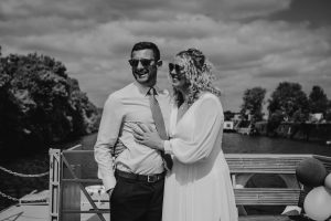 Black and white image of a bride and groom on a boat sailing down the river Thames. The background is sky and water. Bothe of them are wearing sunglasses and smiling.