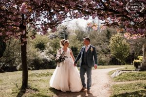 Bride and groom holding hands walking towards the camera. They are surrounded by pink blossom tres. 