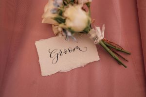 Close up image of a wedding button hole laying on a pink table cloth. The word groom is written in calligraphy on a scrap of water colour paper.