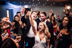Bride on a crowded dance floor jumping with her arm in the air. Surrounded by her guests.