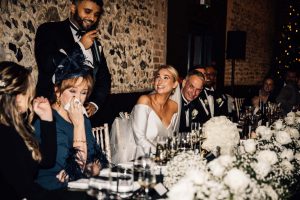 Bride looking towards her husband as he gives a toast. The too table guests are using tissues to wipe their eyes, the bride is smiling.
