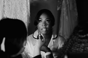 Close u black and white image of a bride having her make up done. Her dress hangs in the background. Taken at a wedding Venue in Leicestershire called Donnington park farm House.