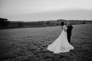 Image of a bride and groom with rolling countryside behind them to showcase Wedding venues in Leicestershire. The image is black and white and the couple are standing in an embrace with her dress fanned out at her feet.