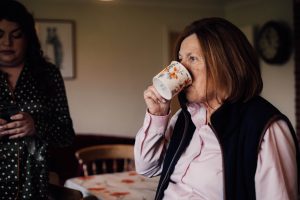 Woman with short brown hair looking out of a window with whilst drinking from a cup. She wears a pink shirt and dark blue Gillet.
