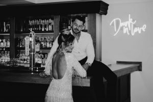 Bride and groom at a bar with a DRUNK IN LVE neon sign hanging on the wall in the background. Brides back is to the camera and the groom is sitting on the bar.