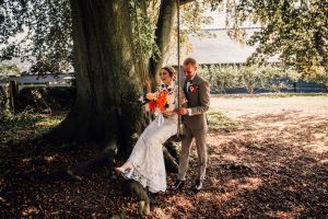 Bride on a swing with the groom pushing her. She is holding her bouquet of orange flowers. Taken in the grounds at a wedding at Sandon Manor.