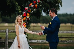 Bride and groom in a field with a flower arch behind them. The groom is holding the brides hand as she smiles into the crowd.