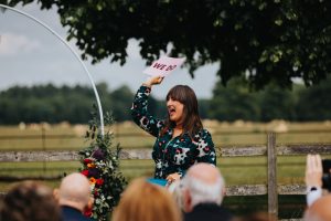 Humanist celebrant wearing green leopard print jumpsuit holding up a sign that says WE DO.