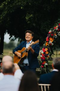 A male singer playing an acoustic guitar while the wedding guests watch on. In the back ground there are bright coloured flowers.